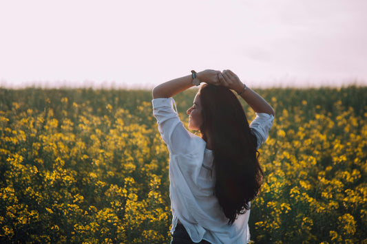 A woman with long brown hair standing in a field of yellow flowers with both her arms over her head smiling and looking to the left.