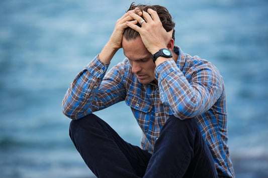 A man wearing a plaid, flannel long sleeve top and blue jean pants, holding in his in frustration sitting with his legs cross with the blue ocean water blurred in the background.