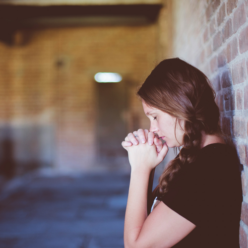 Woman praying to God, asking God for help with her hands clasped together near her face with her eyes closed leaning against a brick wall, blurred background.