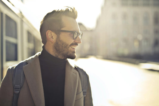 Man smiling with wearing black framed glasses, black turtle neck shirt and a peacoat in the street with blurred buildings in the background. Sunny day.